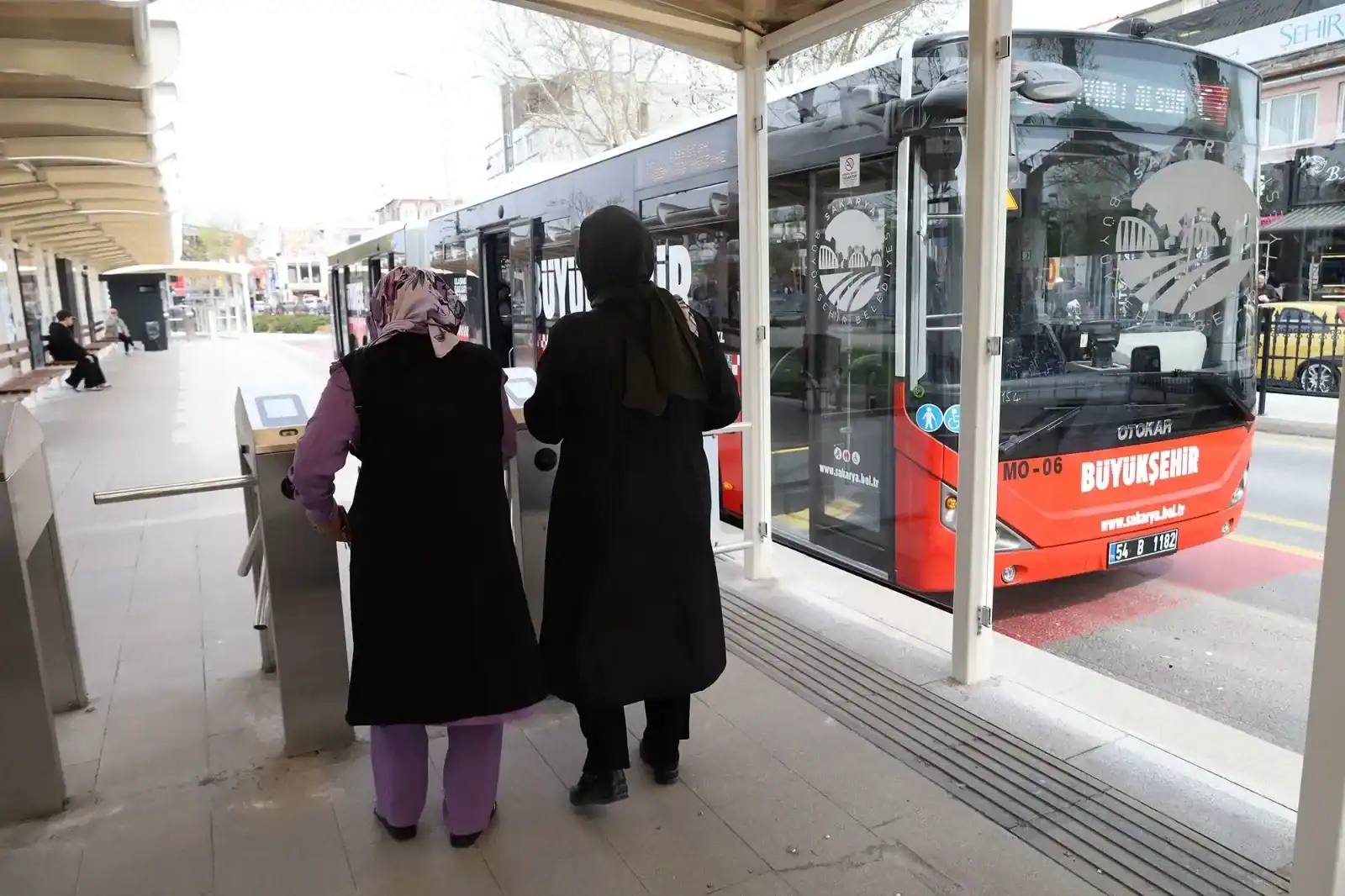 Metrobüs durağında ücretli sistem girişinde iki yolcu, biletlerini alarak bekliyor.